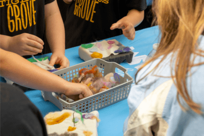 Children engage in a crafting activity at a table. They use colorful wool pieces and felting needles. The mood is focused and creative.
