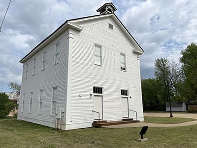 A historical white wooden building with a triangular roof and bell tower on a grassy area. The sky is cloudy, and trees surround the structure.