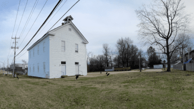 A historical white wooden building with a triangular roof and bell tower on a grassy area. The sky is cloudy, and trees surround the structure.