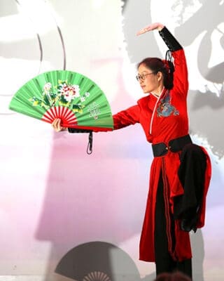 A person in a vibrant red outfit performs a graceful dance holding a green fan with floral patterns, exuding elegance and focus against a soft background.