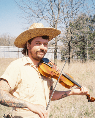 A man with a straw hat and tattoos plays a violin in a sunny field. He smiles warmly, with bare trees and a barn in the background. Calm, rustic vibe.